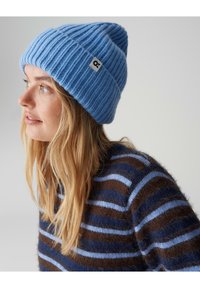 Bonnet en tricot côtelé bleu avec une étiquette, porté avec un pull rayé marron et bleu. Le bonnet est fait d'un tissu doux et texturé.