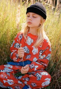 Red sweatshirt and pants with animal prints; model holds a stick, seated in grass. Black knit beanie enhances casual look.