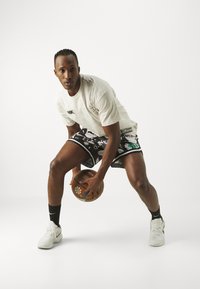 Conjunto deportivo que incluye una camiseta Nike de color claro y textura, y unos pantalones cortos con un patrón en blanco y negro. El jugador sostiene un balón de baloncesto y lleva zapatillas blancas.