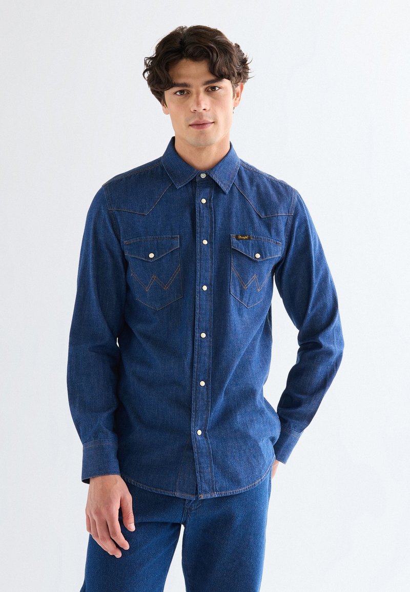 Νεαρός άνδρας κοιτάζει προς τα εμπρός, φοράει σκούρο μπλε τζιν πουκάμισο με κουμπιά τύπου σναπ και τζιν παντελόνι που ταιριάζει, σε φόντο απλού λευκού.