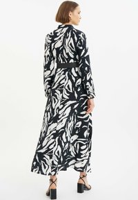 Schwarz-weiß gemustertes langes Kleid mit tailliertem Gürtel, langen Ärmeln und einem luftigen Rock. Strukturierter Stoff mit leichtem Glanz. Blockabsatzsandalen.