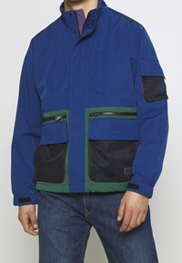 Blå jacka med gröna och svarta meshfickor, dragkedja framtill, hög krage, elastiska manchetter och en avslappnad passform. Har flera förvaringsalternativ.