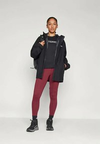 Fekete vízálló dzseki fekete logós pulóver felett, bordó leggings és fekete cipők. Egyszerű dizájn minimális kiegészítőkkel és textúrákkal.