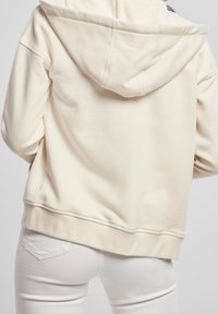 Krémová mikina s kapucí a měkkou texturou, s nastavitelnou šňůrkou, žebrovanými manžetami a spodním lemem, zobrazena zezadu.