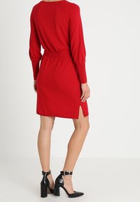 Robe tricotée rouge à manches longues et fente sur le côté, avec une taille cintrée et une texture lisse. Des chaussures à talons hauts noires complètent le look.