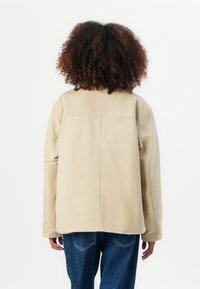 Beige jakke med en afslappet pasform, der har lange ærmer og en central søm. Struktureret stof, kombineret med blå jeans, set fra ryggen.