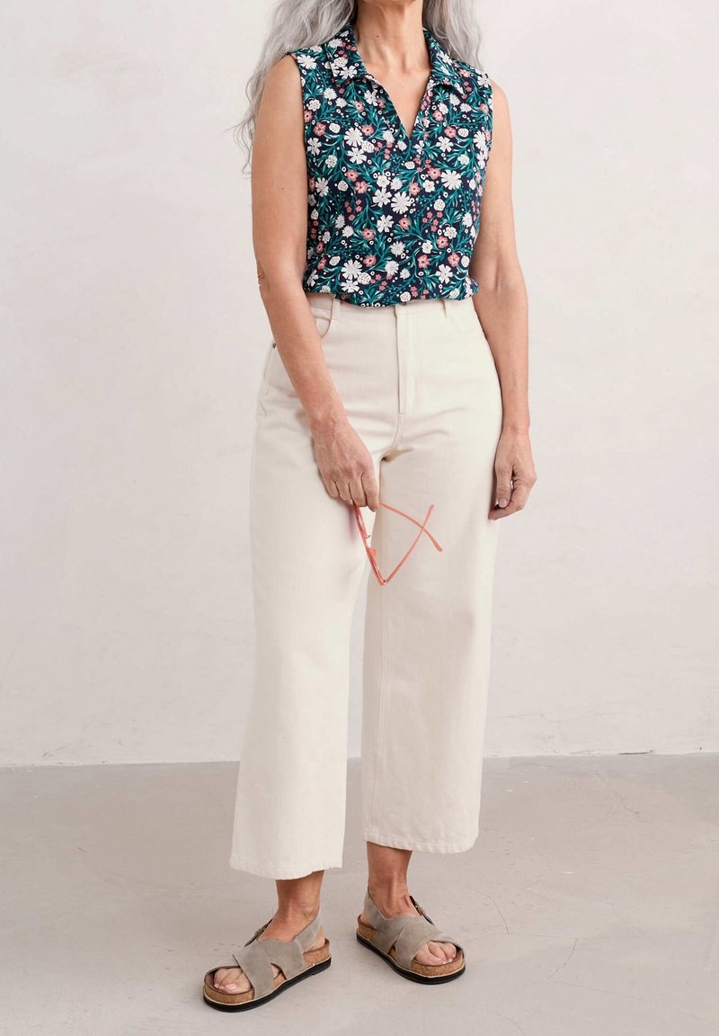 Femme en blouse fleurie sans manches, pantalon large crème, sandales beiges tenant des lunettes à monture rose sur fond uni.