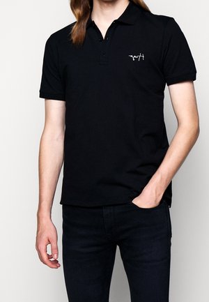 Persona que lleva una camiseta polo negra de manga corta con un pequeño logo blanco en el pecho izquierdo, combinada con pantalones negros, de pie con una mano en el bolsillo.
