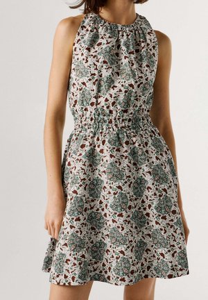 Mouwloze jurk met een geplooide taille en groen-bruin bloemen paisley patroon op lichte stof, gedragen door een staand persoon.