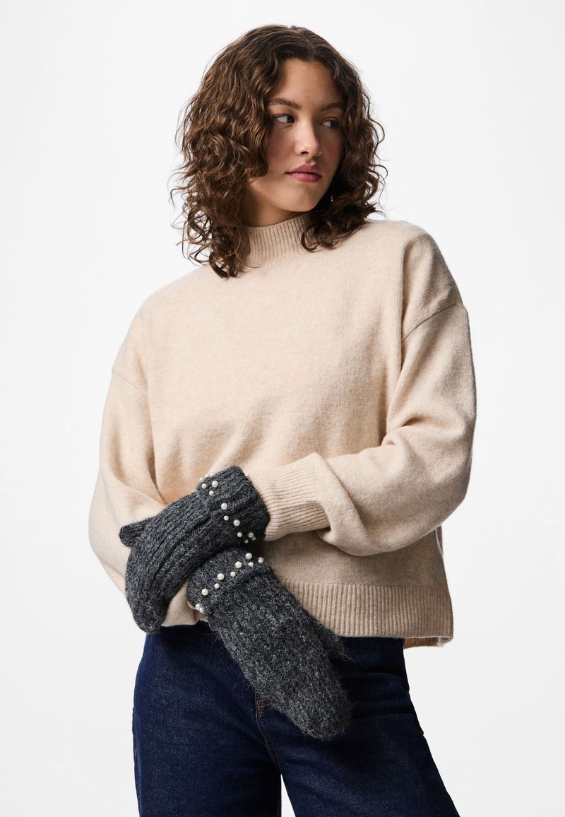 Donna che indossa un maglione beige e jeans scuri, con le mani incrociate che tengono dei guanti di lana grigio scuro decorati con piccoli perline bianche.