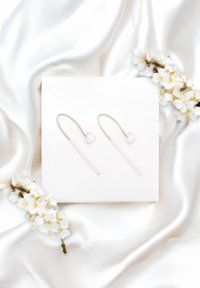 Boucles d'oreilles en argent avec un accent en forme de cœur et des crochets allongés, posées sur un coussin blanc, entourées de fleurs blanches et de satin.