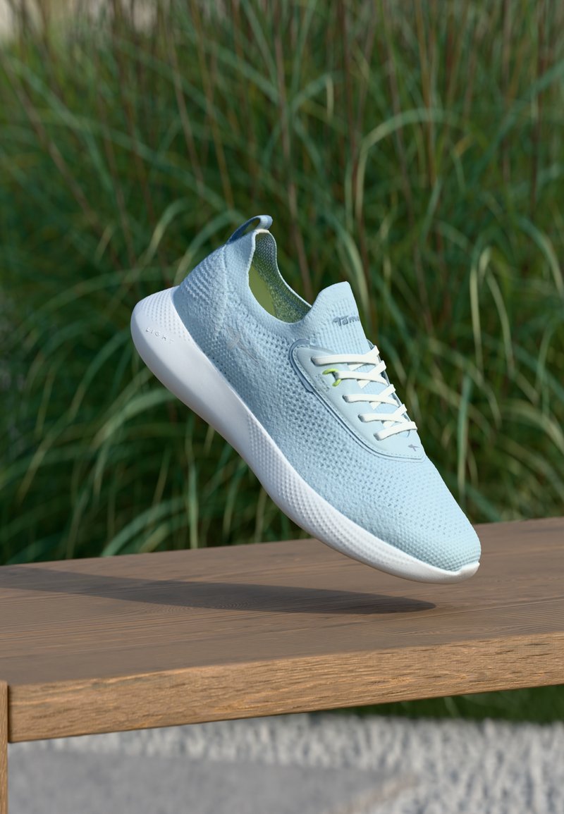 Ανοιχτό μπλε πλεκτό παπούτσι sneaker που πλανάται πάνω από μια ξύλινη παγκάκι με πράσινο χόρτο στο φόντο.