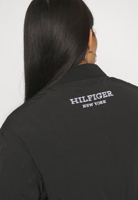 Black bomber jacket featuring ribbed collar, smooth texture, and white embroidered "HILFIGER NEW YORK" logo on the back.