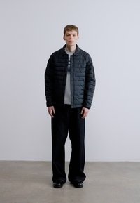 Czarna pikowana kurtka z zapięciem na zatrzaski, noszona na jasnoszarej koszuli i czarnych szerokich spodniach, zestawiona z ciemnymi butami. Gładkie tło.