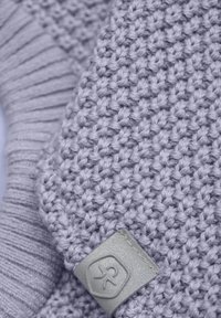 Stickad lavendelfärgad tyg med en texturerad väv, med en ribbad detalj och en grå läderlapp som visar en präglad logotyp.