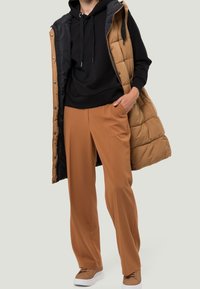 Gevoerde mouwloze vest in bruin over een zwarte hoodie, gecombineerd met losse bruine broeken en lichtbruine sneakers. Soepele texturen en een relaxte pasvorm.