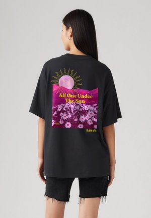 Schwarzes, übergroßes T-Shirt mit einem bunten Grafikdruck eines Sonnen und Blumen auf dem Rücken, mit gelbem Text, der "Alle eins unter der Sonne" sagt.