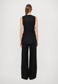 Černá bezrukávová na míru ušitá vesta s detailem zavazování vzadu, v kombinaci s černými širokými kalhotami vyrobenými z texturované látky.
