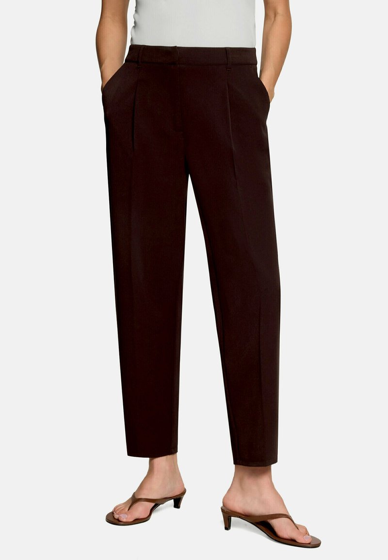 Femme portant un pantalon marron foncé taille haute, fuselé, avec des poches latérales et des sandales marron à talons avec lanière entre les orteils.