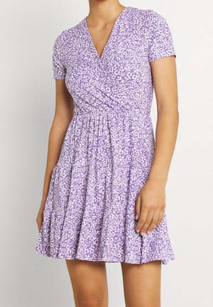 Vrouw draagt een paarse bloemen wikkeljurk met korte mouwen en een geplooide, gelaagde rok tegen een effen lichte achtergrond.