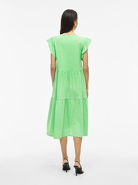 Zielona, warstwowa midi sukienka z krótkimi, falbaniastymi rękawami, miękkim materiałem i okrągłym dekoltem. Noszona z czarnymi sandałami na obcasie.