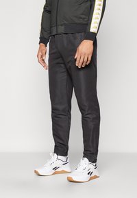 Black athletic pants with elastic cuffs, featuring a smooth texture and a side panel with a logo. Worn with white sneakers.