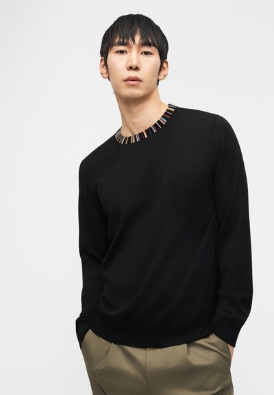 Jeune homme portant un pull noir à manches longues avec un col rayé multicolore et un pantalon beige, debout devant un fond clair uni.