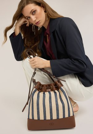 Junge Frau in marineblauem Blazer und weißer Hose hält eine gestreifte, gewebte Henkeltasche mit Lederakzenten und sitzt vor einem einfarbigen Hintergrund.