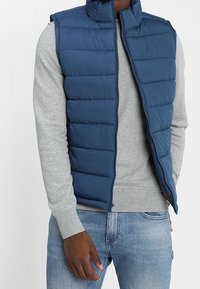 Marinblå vadderad väst med hög krage och dragkedja, bärs över en grå långärmad skjorta. Tyget har en slät yta och kviltat mönster.