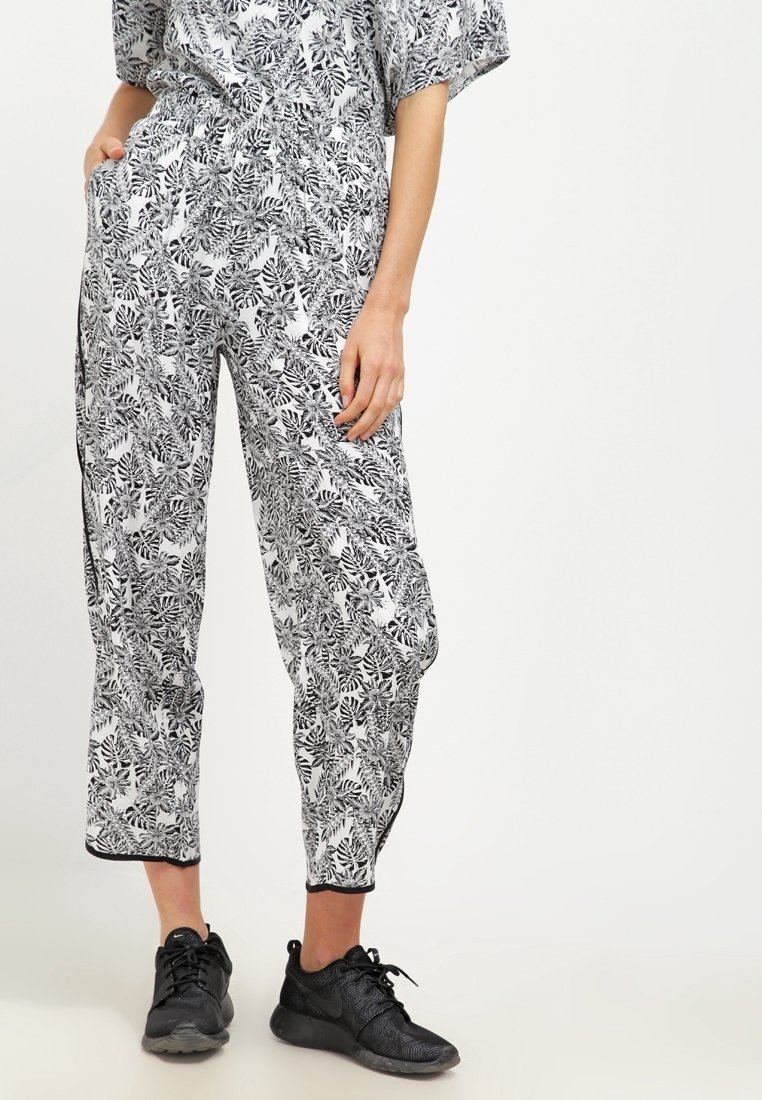 Personne portant un pantalon à motifs feuilles noir et blanc et un haut assorti, associé à des baskets de sport noires, se tenant avec une main dans la poche.
