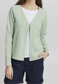 Ljusgrön zip-up kofta med ribbad textur, raglanärmar och v-ringad design, bärs över en vit skjorta och marinblå shorts.