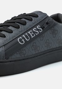 Adidași dintr-un material texturat gri închis, cu o talpă din cauciuc negru. Prezintă șireturi negre și un fermoar lateral cu logo-ul "GUESS" în alb.