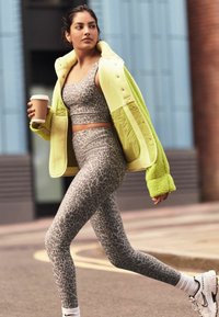 Neon gele oversized jas met een pluche textuur, grijs sportbeha met luipaardpatroon, bijpassende legging en witte sneakers, terwijl ze een koffiepot vasthoudt.
