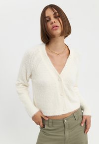 Witte, pluizige, korte cardigan met een diepe V-hals en vijf knopen. Geribde manchetten en zoom voegen textuur toe. Draag over kaki broek.