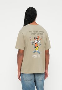 Camiseta verde oliva con un gráfico de Mickey Mouse esquiando, con el texto "EL ARTE DEL ESQUÍ" y detalles coloridos. Ajuste casual y holgado.