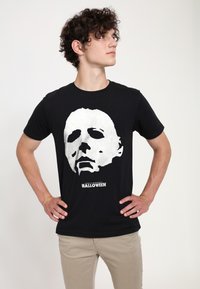 Černé bavlněné tričko s velkým bílým grafickým znakem tváře masky a textem "JOHN CARPENTER'S HALLOWEEN" pod ním. Krátké rukávy, kulatý výstřih.