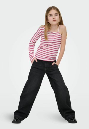 Fată tânără cu păr lung, purtând un top cu o singură mânecă în dungi și pantaloni negri evazați, stând cu mâinile în buzunare, pe un fundal simplu.