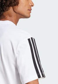 Biely tričko s čiernymi trojprúžkami na rukávoch. Vyrobené z bavlny s jemnou textúrou, s klasickým dizajnom okrúhleho výstrihu.