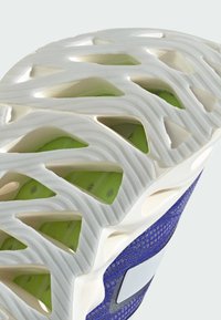 La suola della scarpa presenta una base in gomma bianca testurizzata con inserti di ammortizzazione verdi e un intricato design a reticolo, completata da una tomaia viola.