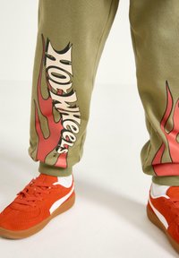 Olivengrønne svedbukser med røde flammegrafikker og "Hot Wheels"-tekst; parret med klare orange sneakers med hvide accenter og gummisåler.