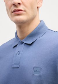 Gaiši zils polo krekls ar teksturētu virsmu, trīs pogu aizdare un tonāls "BOSS" logo uz krūšu daļas.