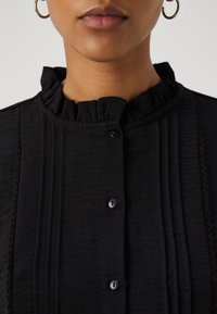 Blusa negra con cuello volante, pliegues verticales y costuras decorativas, con botones negros en la parte delantera. Textura suave.