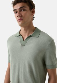 Lys grøn polo-skjorte med V-hals, korte ærmer, struktureret strikmønster og ribbet krave og manchetter. Tætsiddende design.