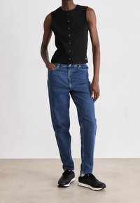 Schwarzes ärmelloses Oberteil mit Knöpfen vorne, kombiniert mit locker sitzender blauer Jeans. Model trägt schwarze Sneakers. Neutraler Hintergrund.