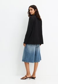 Čierna sako s priliehavým strihom, nosené cez voľné denimové culottes. Look je doplnený hnedými sandálmi na remienky, na bielom pozadí.