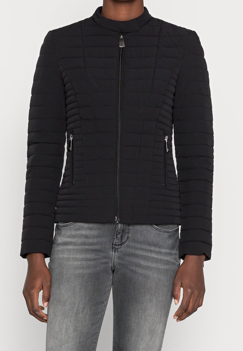 Personne portant une veste noire matelassée ajustée avec fermeture éclair sur le devant et poches latérales zippées, associée à un jean gris délavé.
