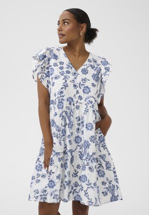 Žena v bielych šatách s modrým kvetinovým vzorom, s volánikovými rukávmi a výstrihom do V, stojaca s jednou rukou v vrecku, pozerajúca sa do strany.