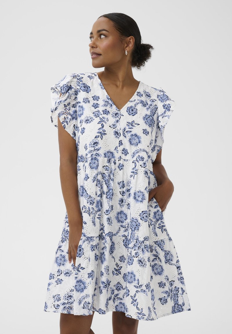Femme portant une robe blanche à imprimé floral bleu, avec des manches à volants et un col en V, debout avec une main dans la poche, regardant sur le côté.