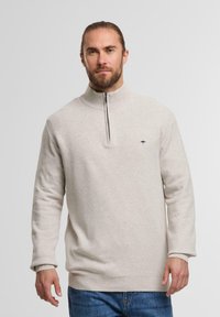 Mann mit Bart, der einen beigen Reißverschluss-Pullover und blaue Jeans trägt, steht vor einem hellen, einfarbigen Hintergrund.