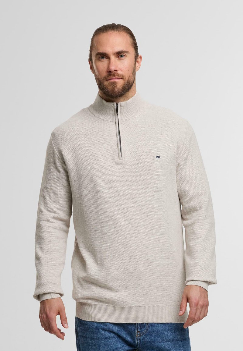 Mann mit Bart, der einen beigen Reißverschluss-Pullover und blaue Jeans trägt, steht vor einem hellen, einfarbigen Hintergrund.